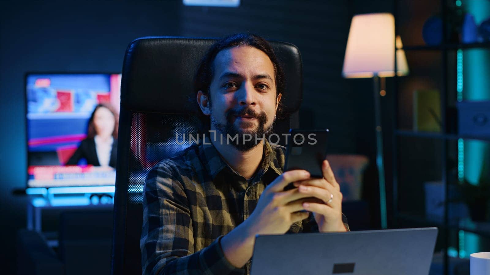 Man relaxing in apartment sitting at office desk, texting friends on cellphone, enjoying leisure time. Smiling remote worker taking break to talk with mates on internet using phone, camera A