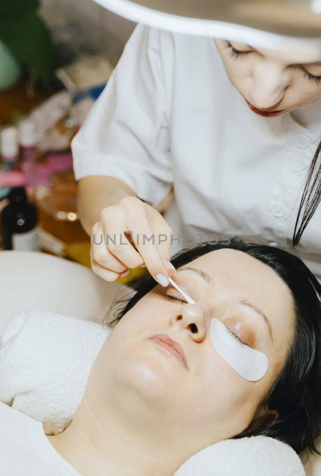 Portrait of a young beautiful Caucasian brunette cosmetologist girl in a white coat who degreases eyelashes on the lower eyelid with a cotton swab to a woman lying on a cosmetology table in a home beauty salon, close-up side view.