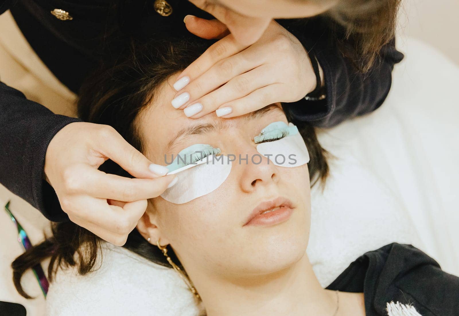 Portrait of a young unrecognizable Caucasian girl cosmetologist in a dark jacket, who cleans the remnants of products from the eyelashes on the silicone roller of the left eye using a cotton swab to a teenage client lying on the cosmetology table in a home beauty salon for eyelash lifting, top side close-up view with selective focus.