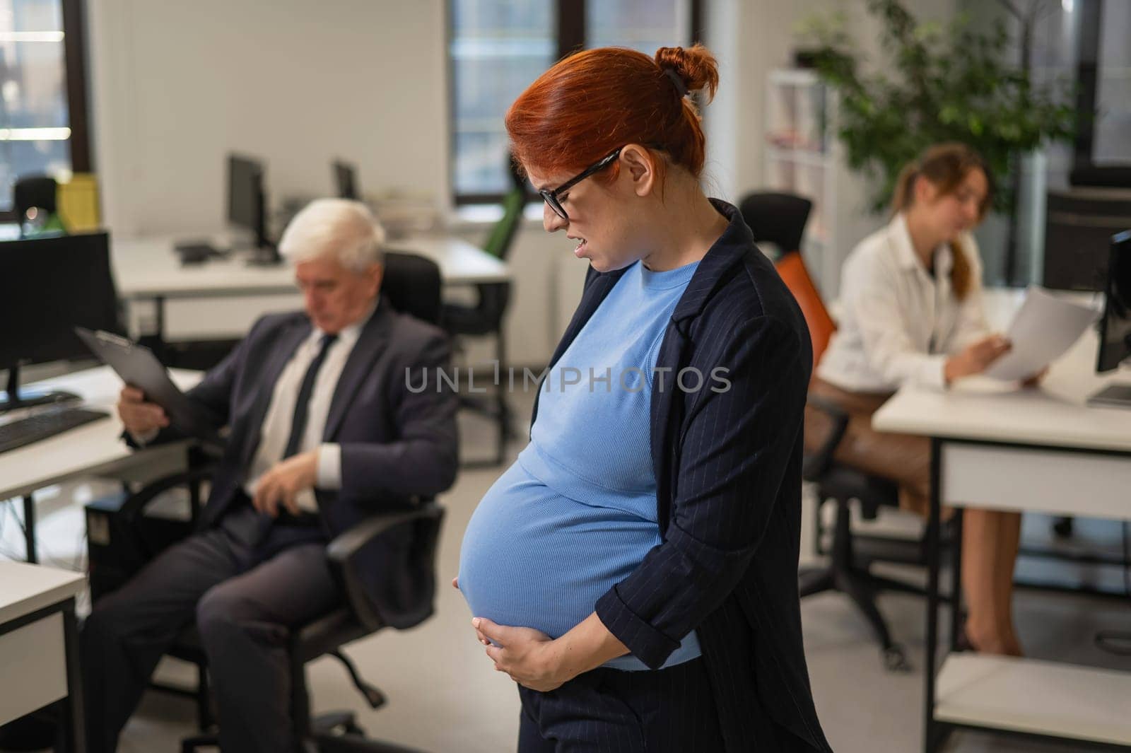 A pregnant woman suffers from pain and holds her stomach while standing in the middle of the office next to her colleagues. by mrwed54