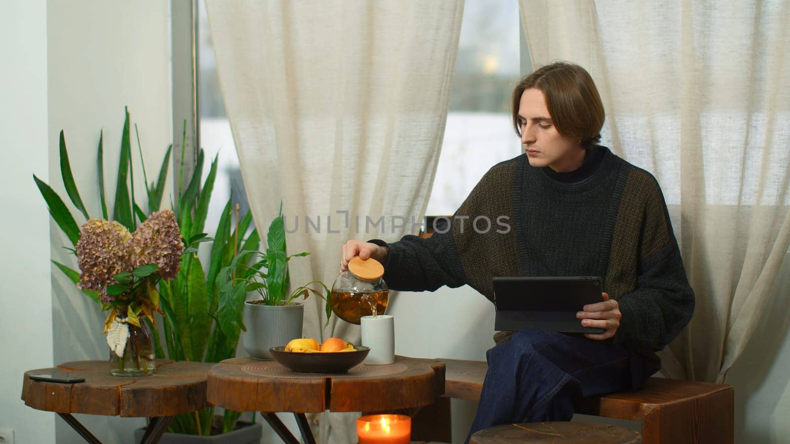 Young man is sitting in cafe with tea and tablet. Media. Man in cozy cafe is working on tablet and drinking tea. Young freelancer works in cafe with tablet.