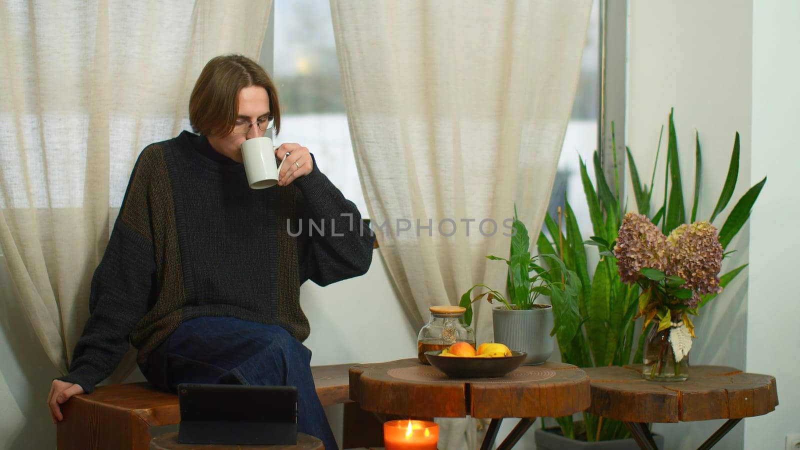 Handsome student drinks tea and looks at tablet in cafe. Media. Young man with glasses is watching lecture on tablet in cafe. Young man is relaxing and studying in cozy cafe.
