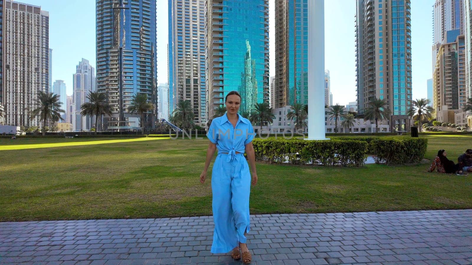 Happy woman in blue summer suit walking in city park. Action. Green meadow and skyscrapers on the background