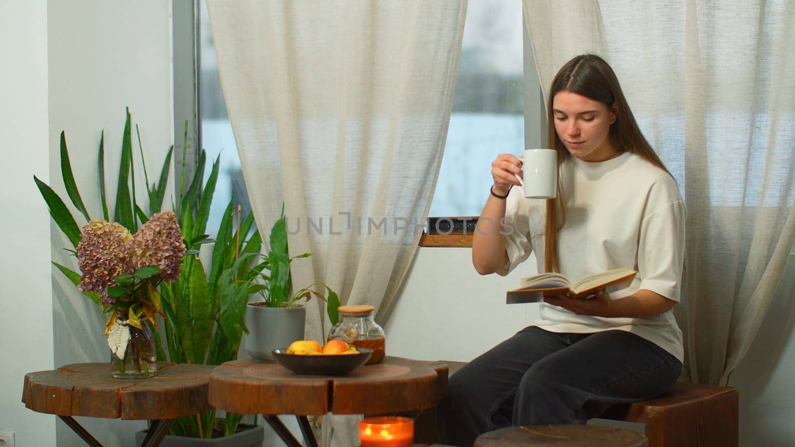 Young woman is reading book with tea in cozy cafe. Media. Beautiful young woman is reading book in cafe. Cozy winter day in cafe with mug of tea and favorite book.