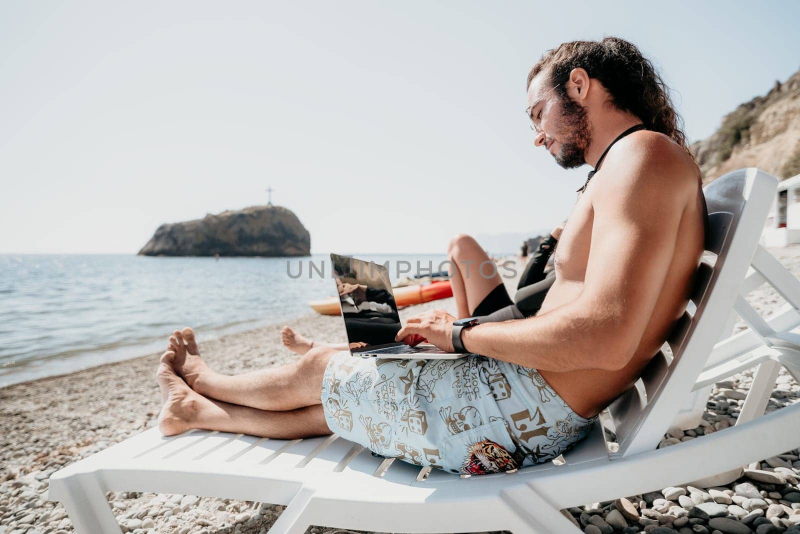 Digital nomad, Business man working on laptop by the sea. Man typing on computer by the sea at sunset, makes a business transaction online from a distance. Freelance, remote work on vacation