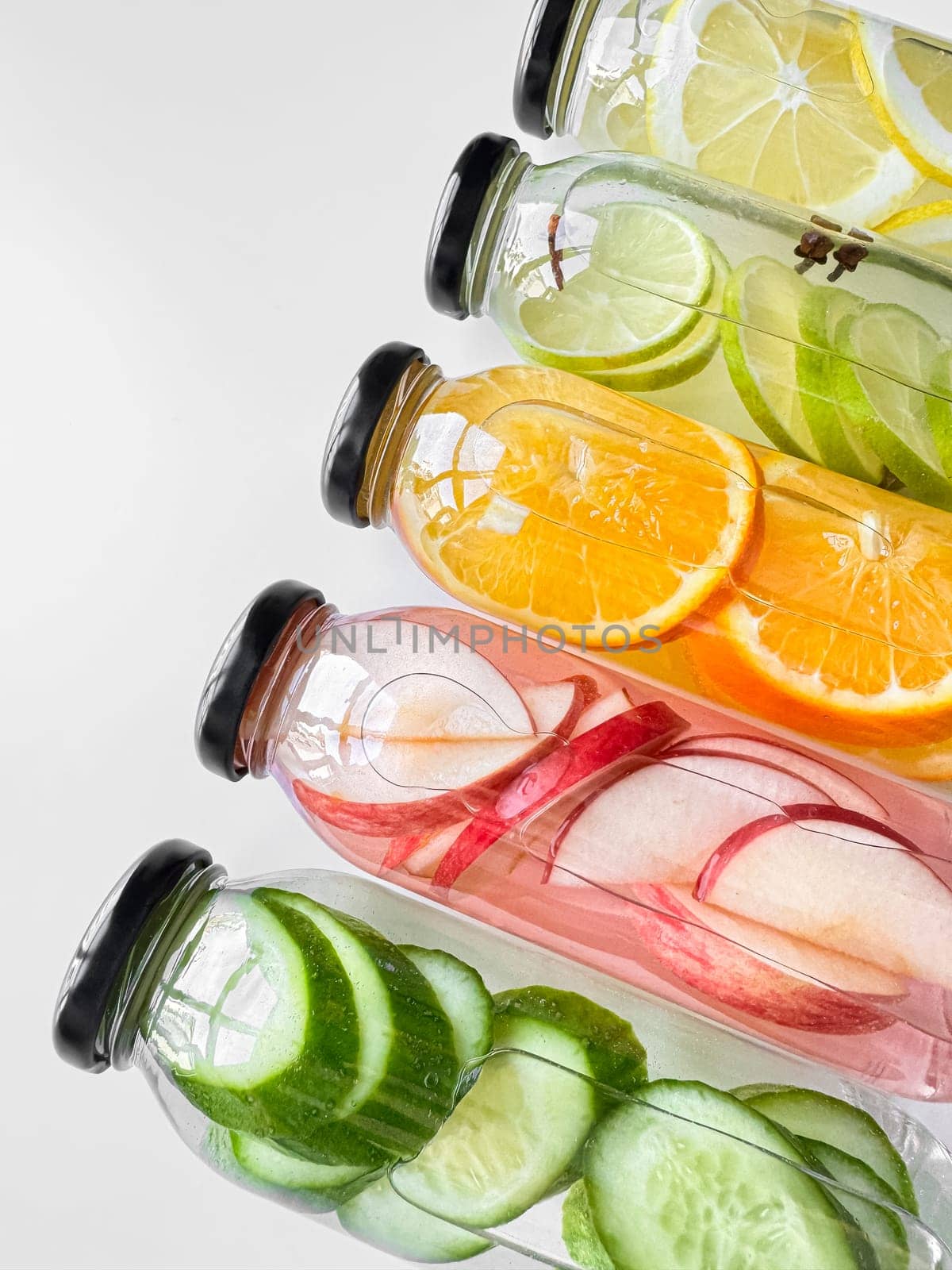 Stacked glass bottles with detox drinks with cucumber, apple, orange, and lemon slices on white background. Vertical orientation, refreshing beverage concept for design and print. High quality photo