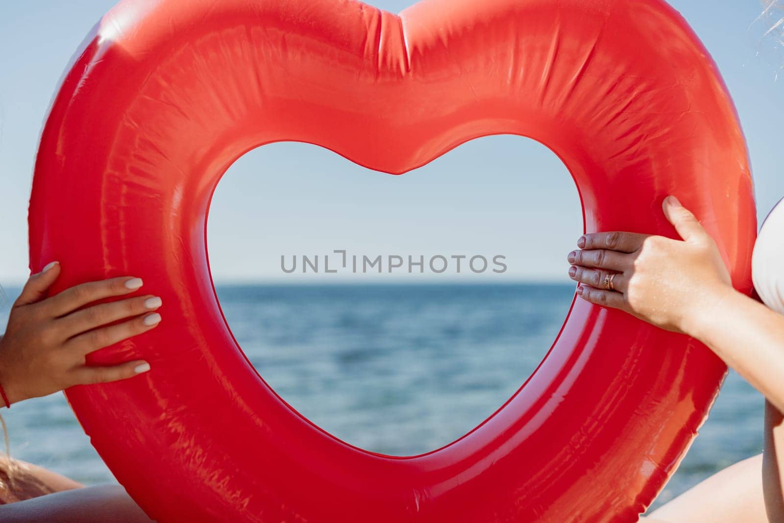 Two women are holding a red inflatable heart on the beach. The heart is open, revealing the ocean behind it. Concept of love and happiness