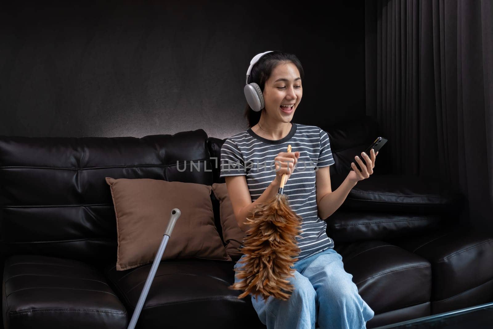 Smiling woman dusting with feather duster and listening to music on headphones. Concept of multitasking and joyful housework.