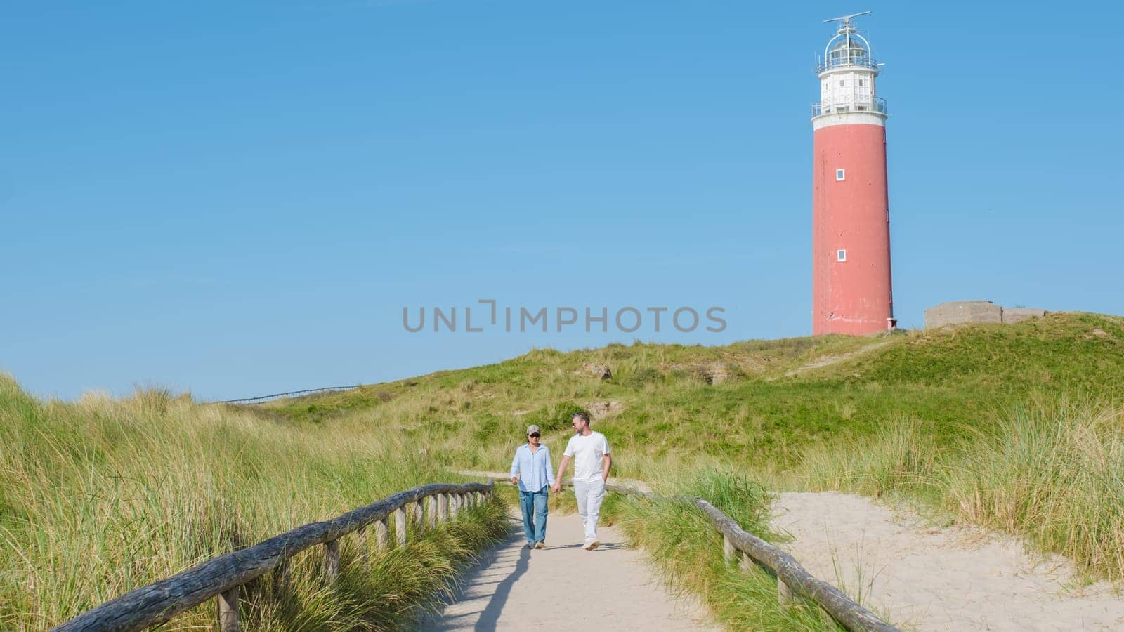 A man and a woman are strolling leisurely along a winding path near the iconic Texel lighthouse, enjoying the scenic view of the surrounding landscape. The iconic red lighthouse of Texel Netherlands