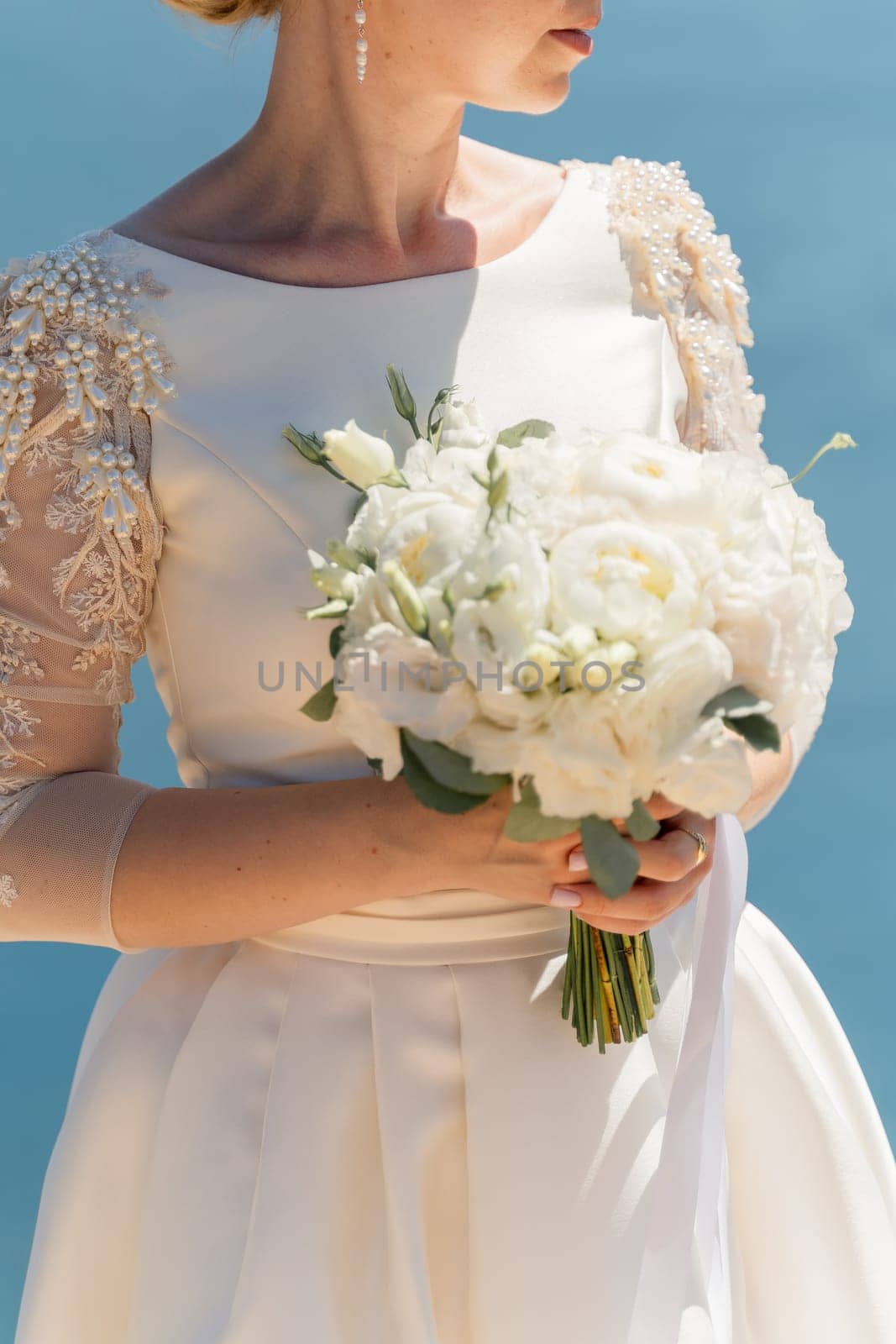 bride is holding a bouquet of white flowers. The bouquet is large and has a lot of flowers in it. The woman is wearing a white dress and a gold ring on her finger. Scene is elegant and romantic