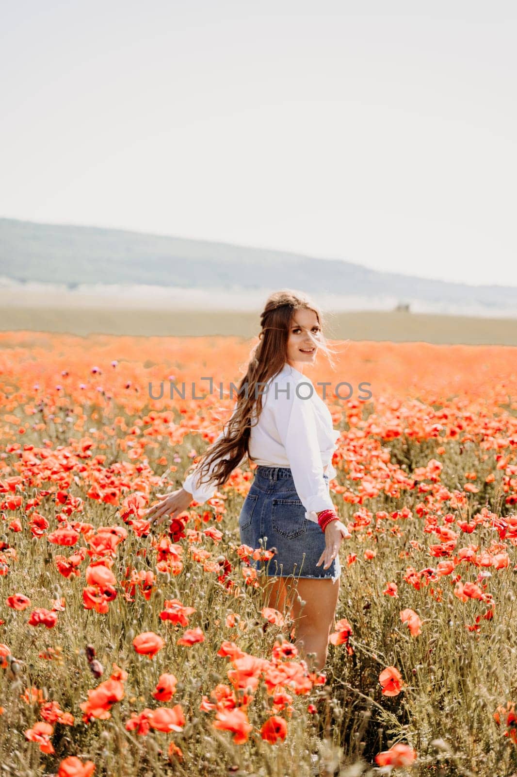 Happy woman in a poppy field in a white shirt and denim skirt with a wreath of poppies on her head posing and enjoying the poppy field