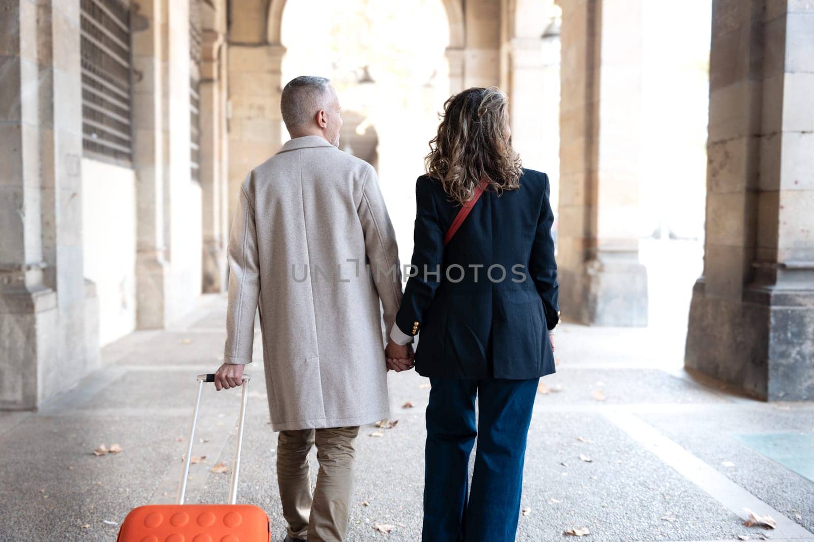 Smiling middle-aged Caucasian tourist couple walking hand-in-hand carefree with their luggage looking at each other. Happy husband and wife enjoying their outdoor vacation on a sunny winter day