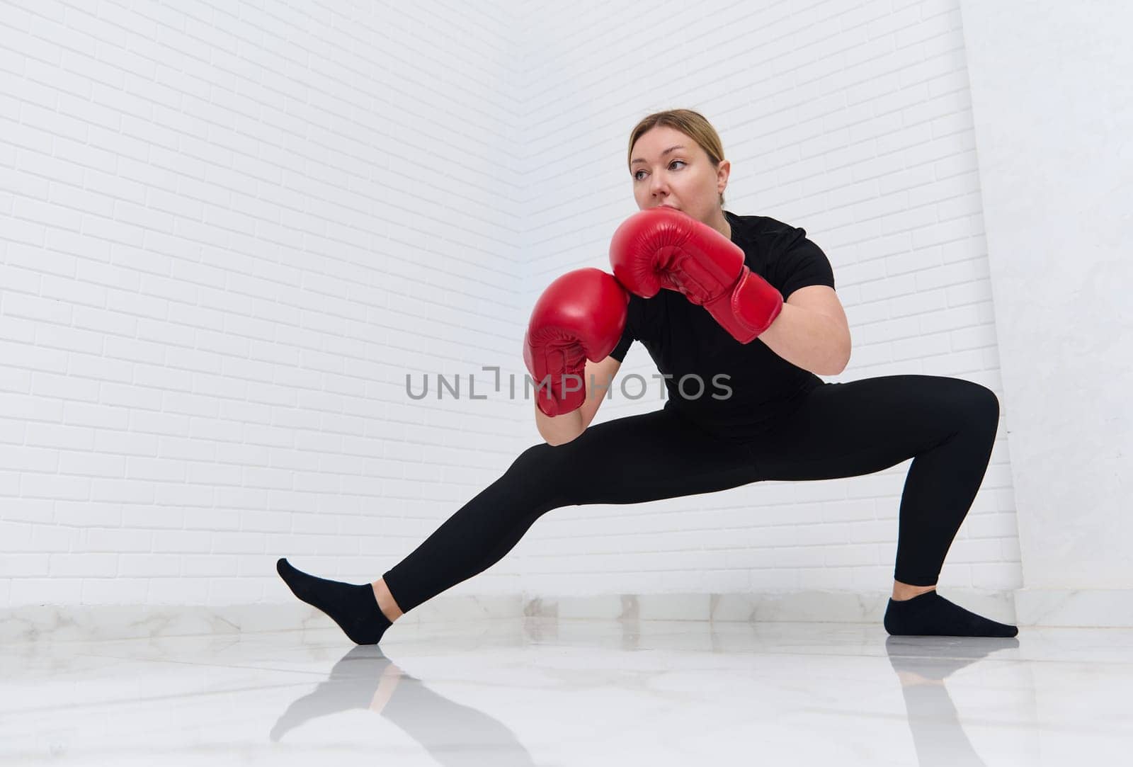 European woman fighter boxer in red boxing gloves, doing stretching exercises on legs,warming up, getting for combat, match, challenge, isolated over white background. Martial art. People and sport.