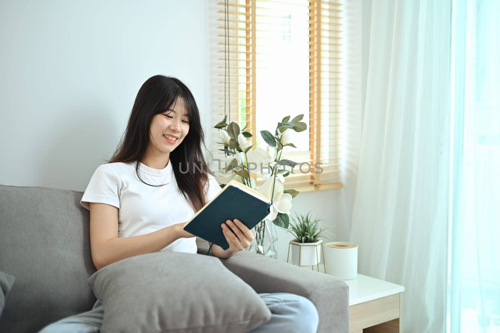 Happy young woman wearing casual clothes relaxing in her living room and reading book.