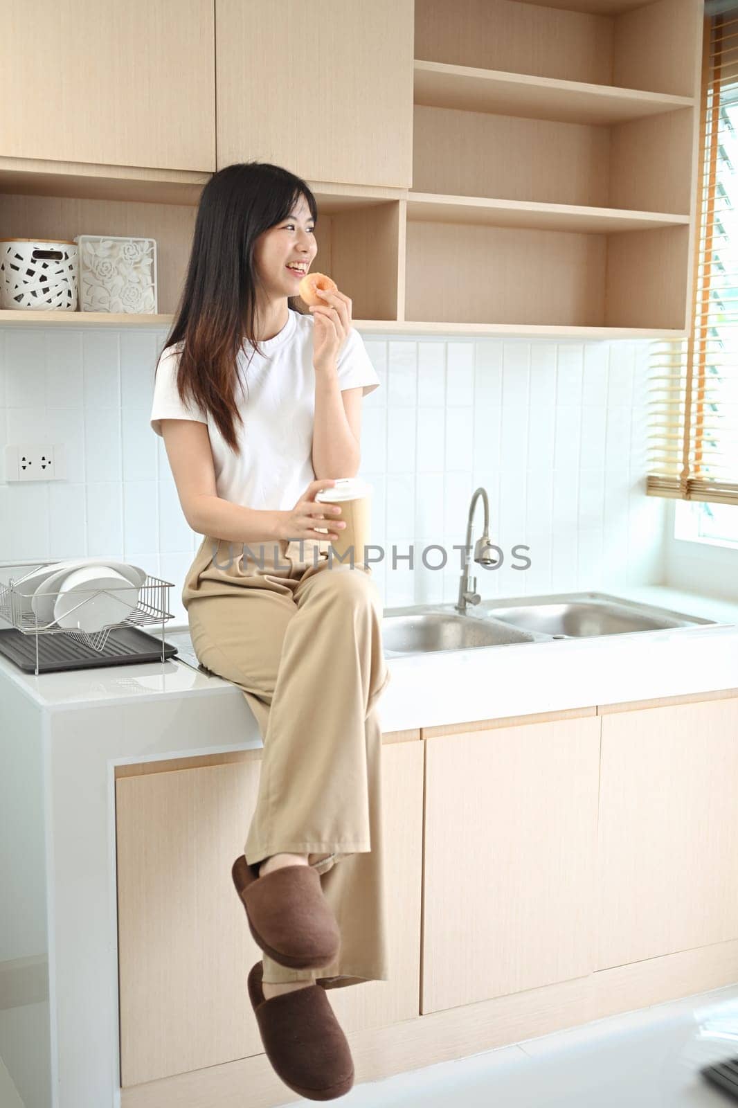 Portrait of smiling casual young woman eating a donut and drinking coffee in the kitchen.