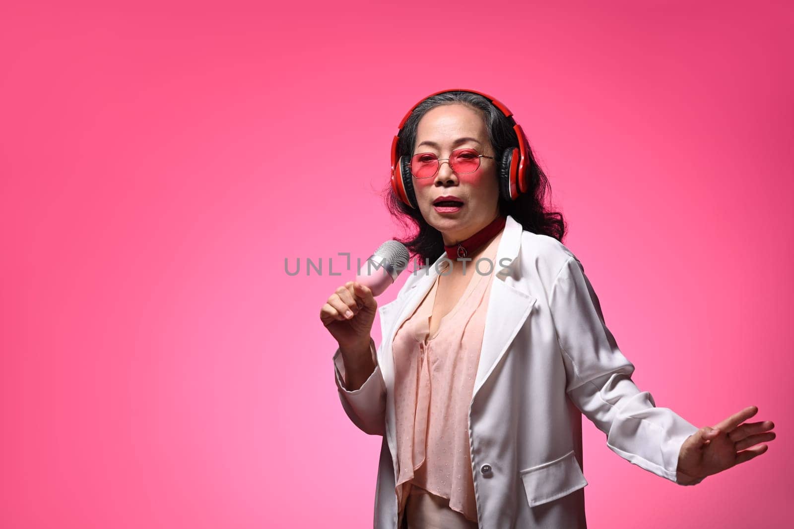 Stylish middle age lady in headphone singing with microphone on pink background.