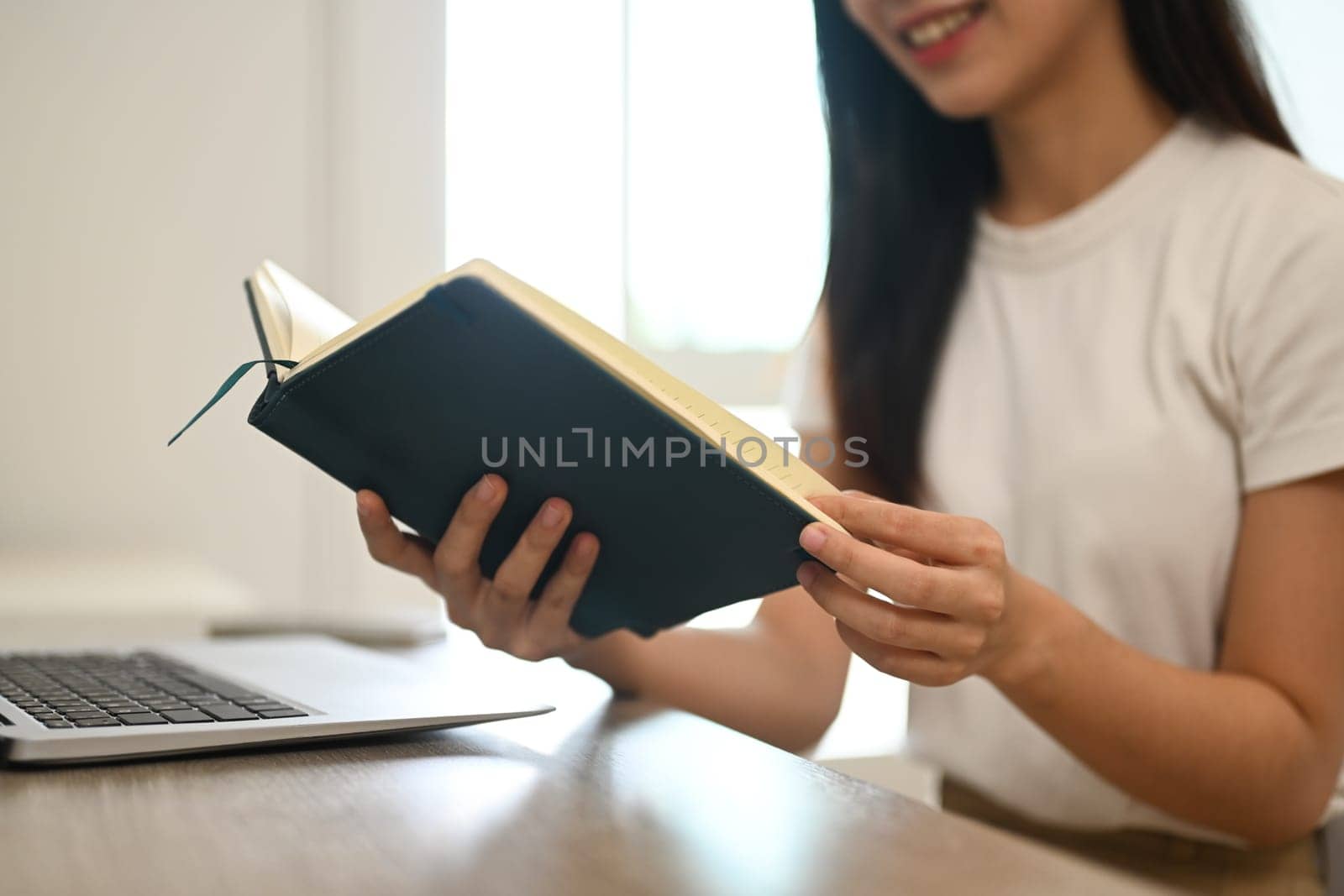 Peaceful young Asian woman in casual clothes reading book on desk at home.