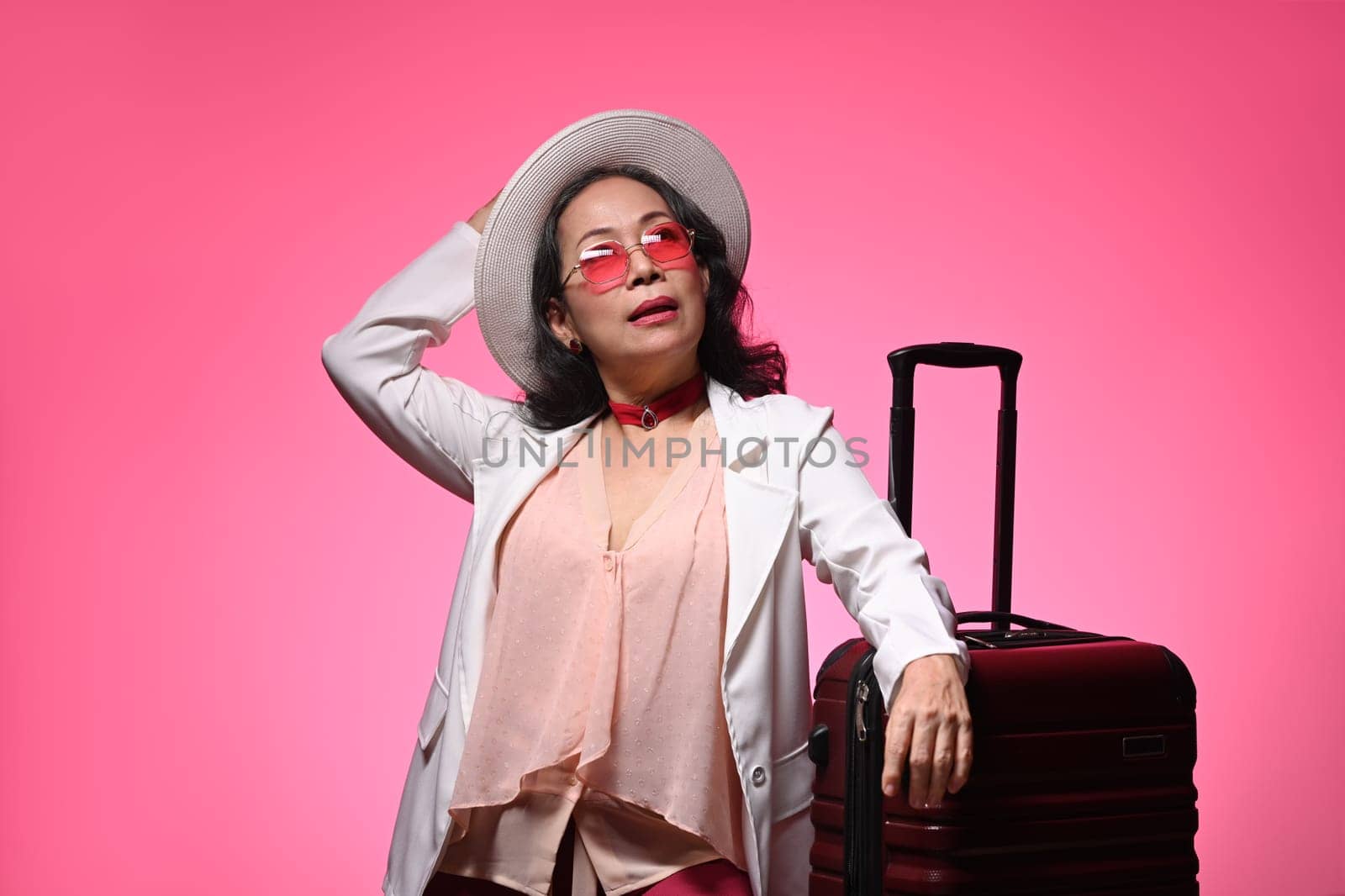 Confident middle age woman in sunglasses and straw hat standing with suitcase on pink background by prathanchorruangsak