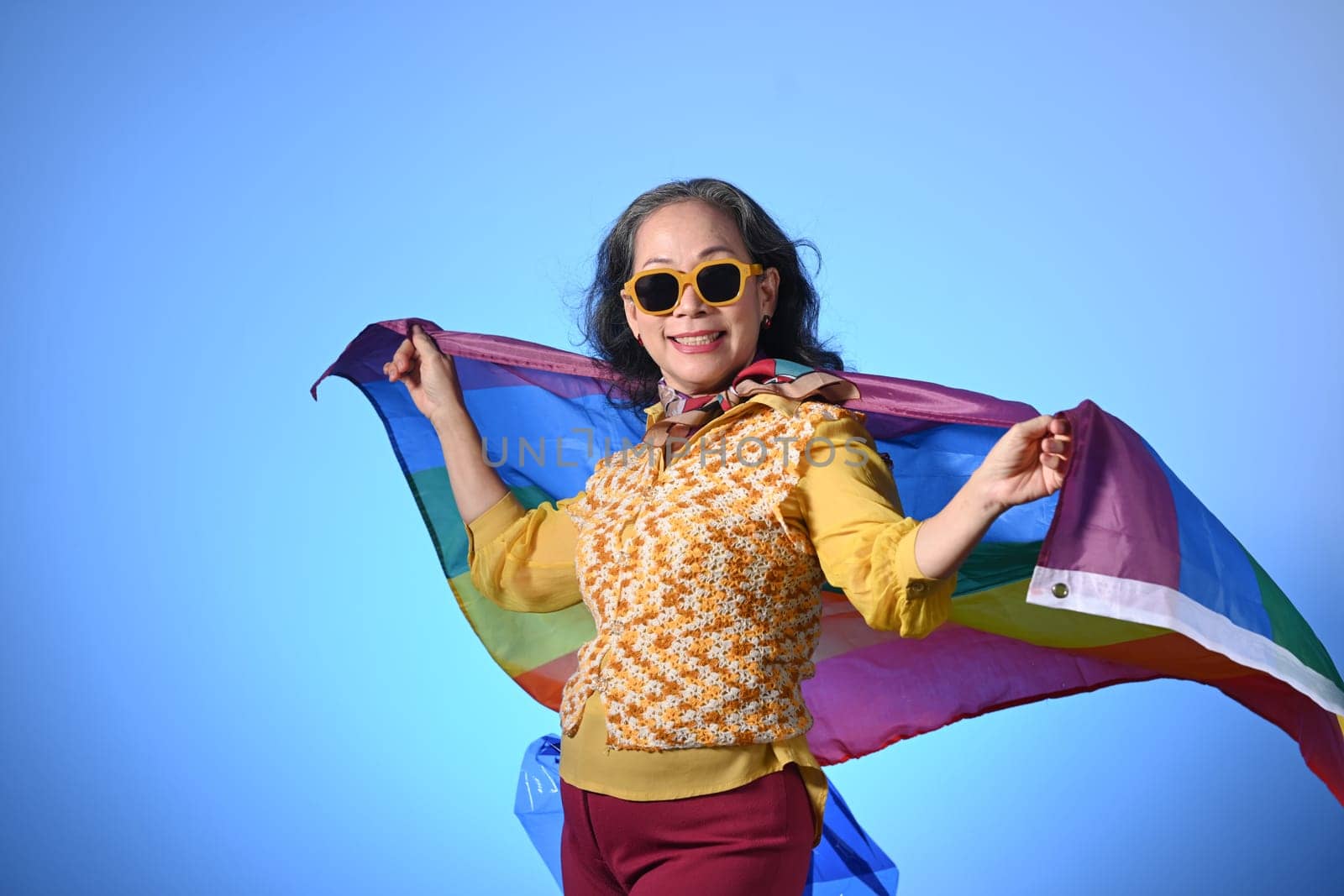 Older lady holding a rainbow flag on blue background LGBT, human rights, freedom and equality concept.