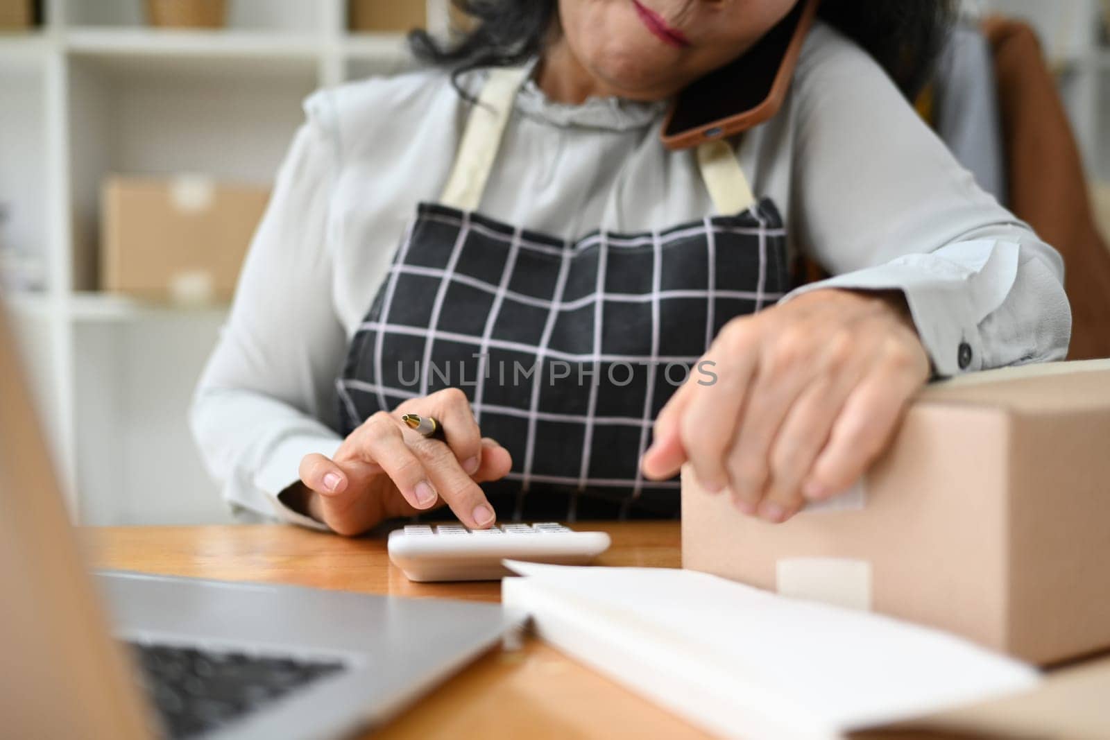 Middle age female small business owner having phone conversation and calculating expense at desk.