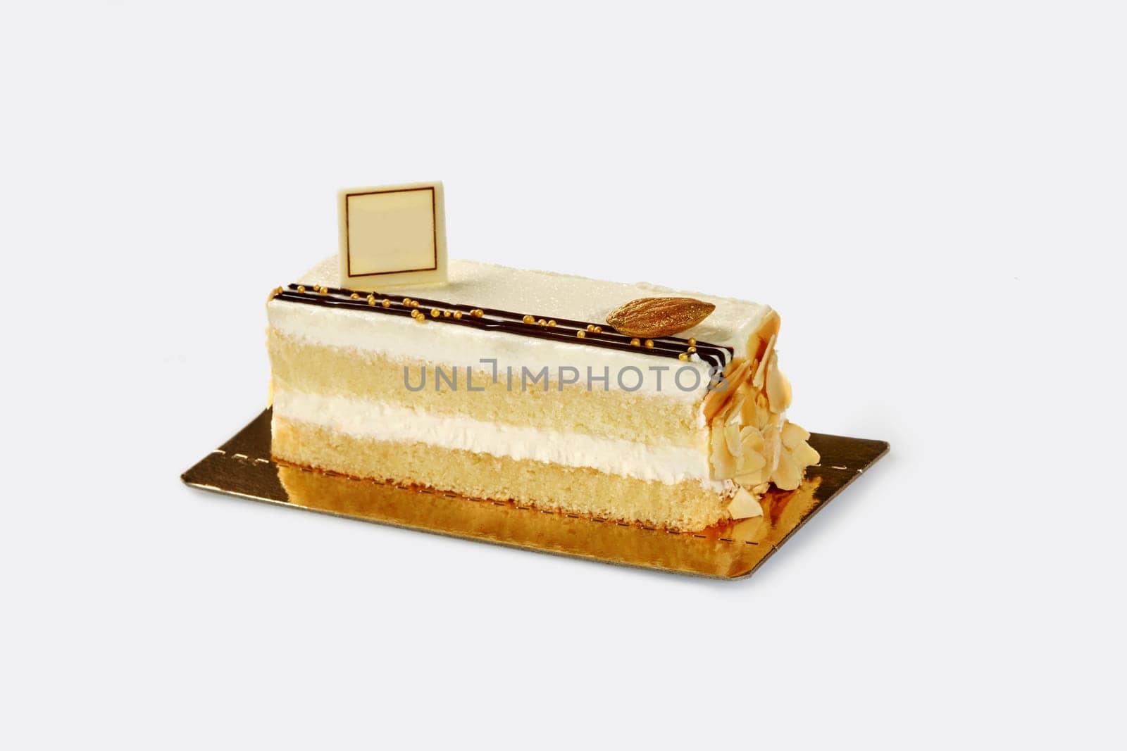 Slice of delectable airy fluffy biscuit cake with delicate light cream decorated with gilded caramelized almond and flakes, presented on gold cardboard against white backdrop