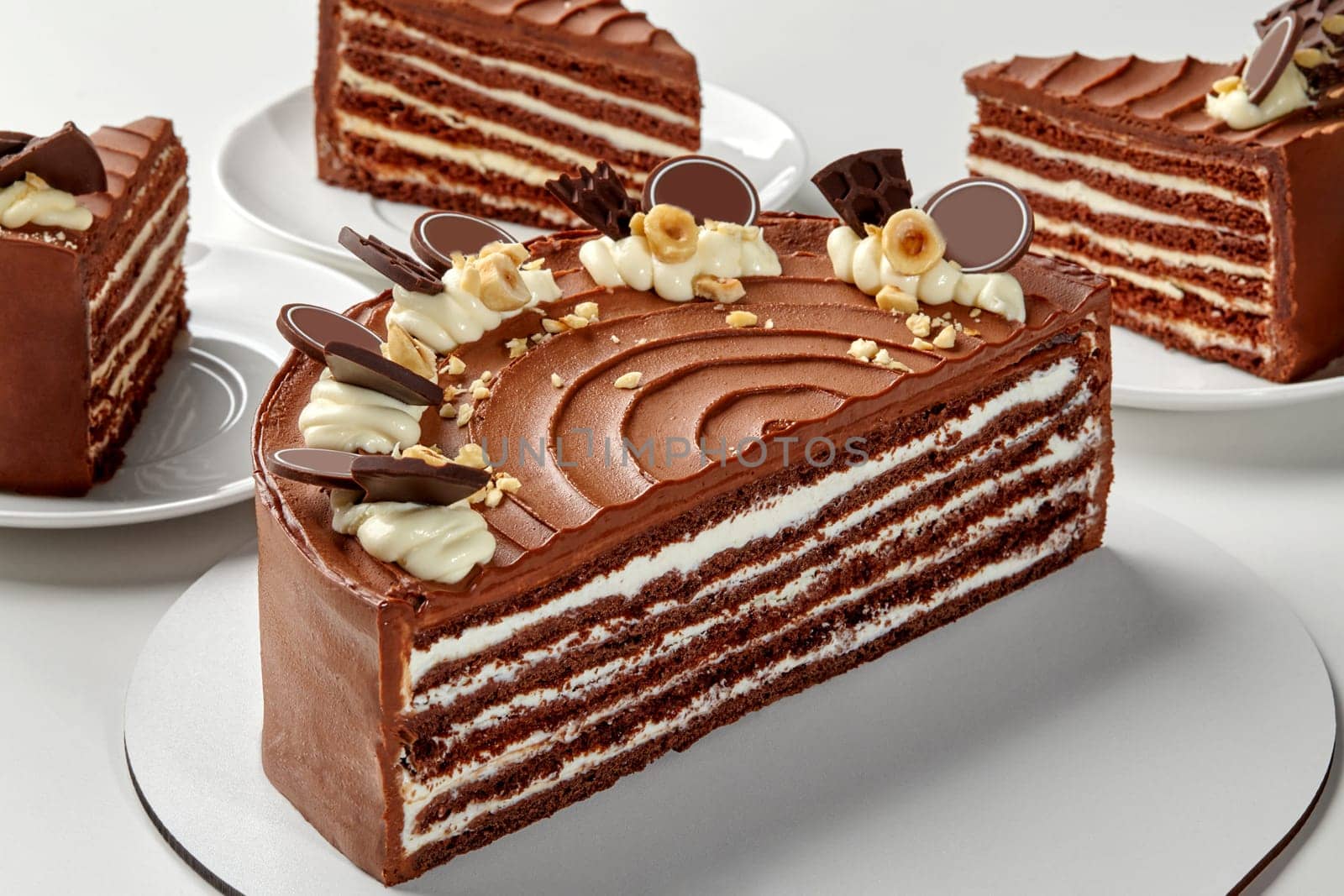 Sliced decadent layered cake boasting alternating layers of honey chocolate sponge and whipped sour cream, adorned with cream swirls, hazelnut pieces, and dark chocolate accents on white background