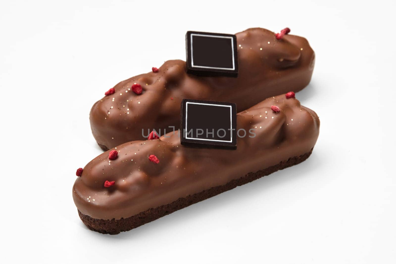 Two streusel-based bars topped with pecans, coated in milk chocolate, adorned with dried berries and dark chocolate brand plaque, on white background. Artisan handmade sweets