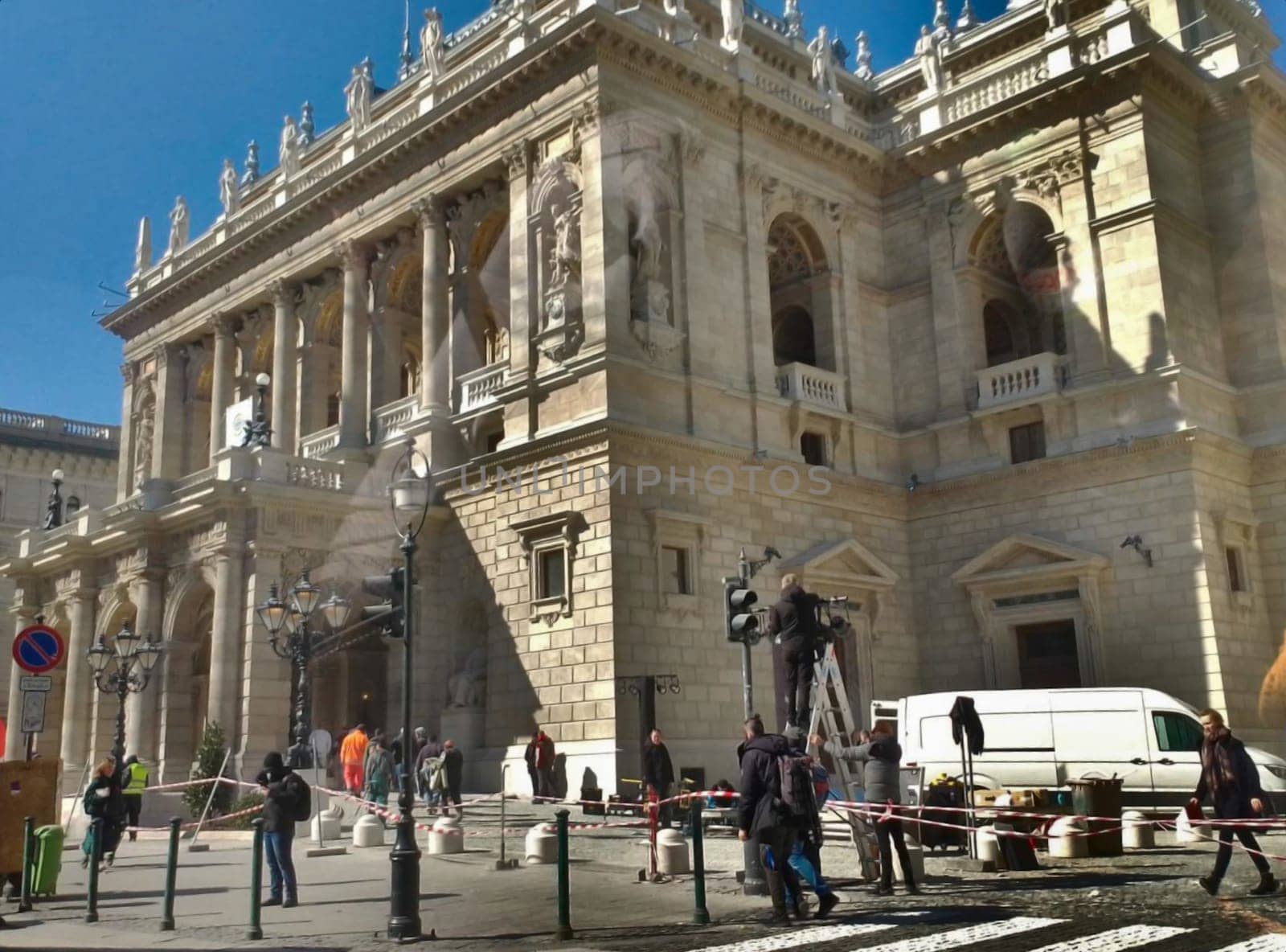 Road services are repairing a traffic light, against the backdrop of a building with columns and statues.