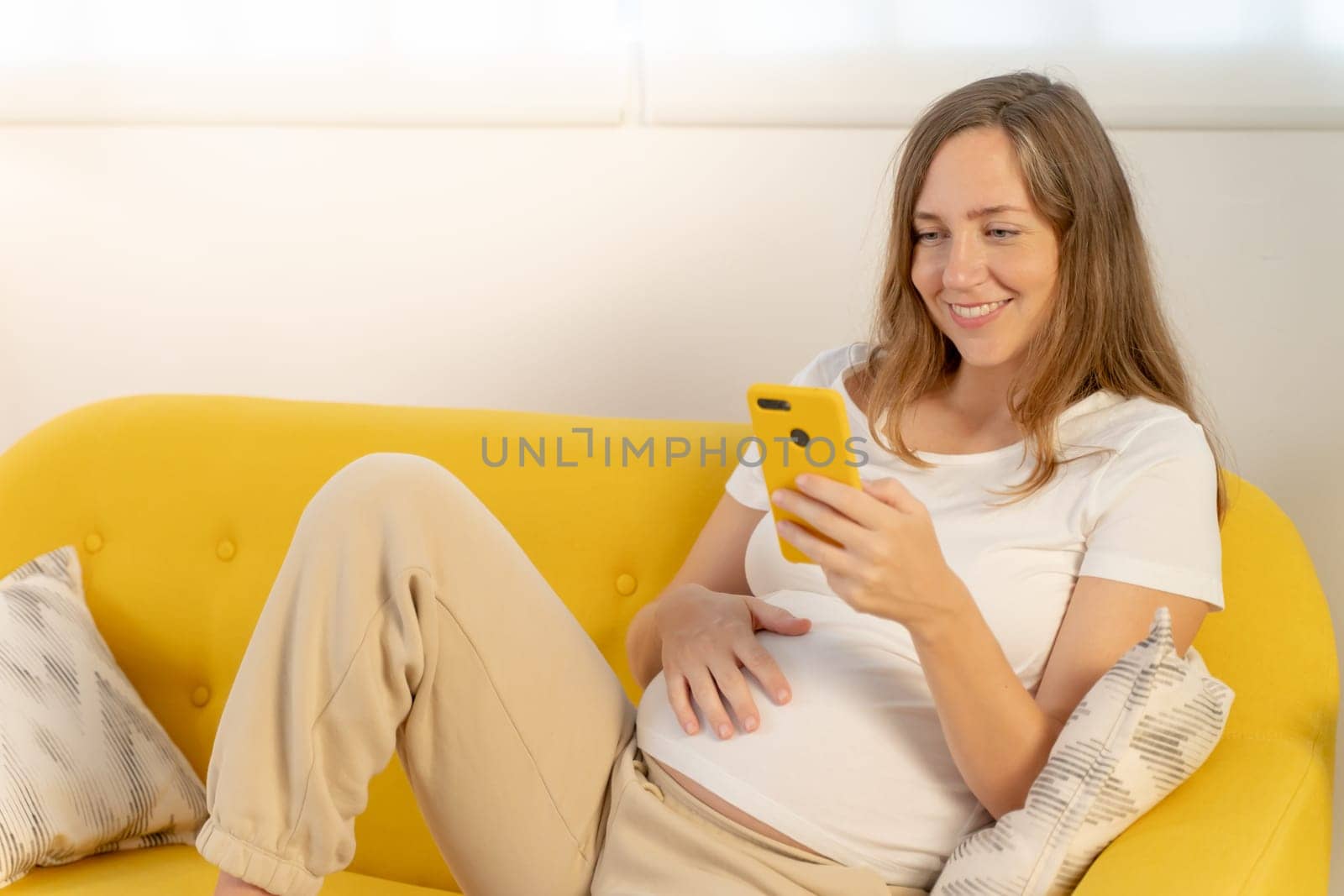 Pregnant woman enjoying leisure time on comfy yellow couch, looking phone. High quality photo