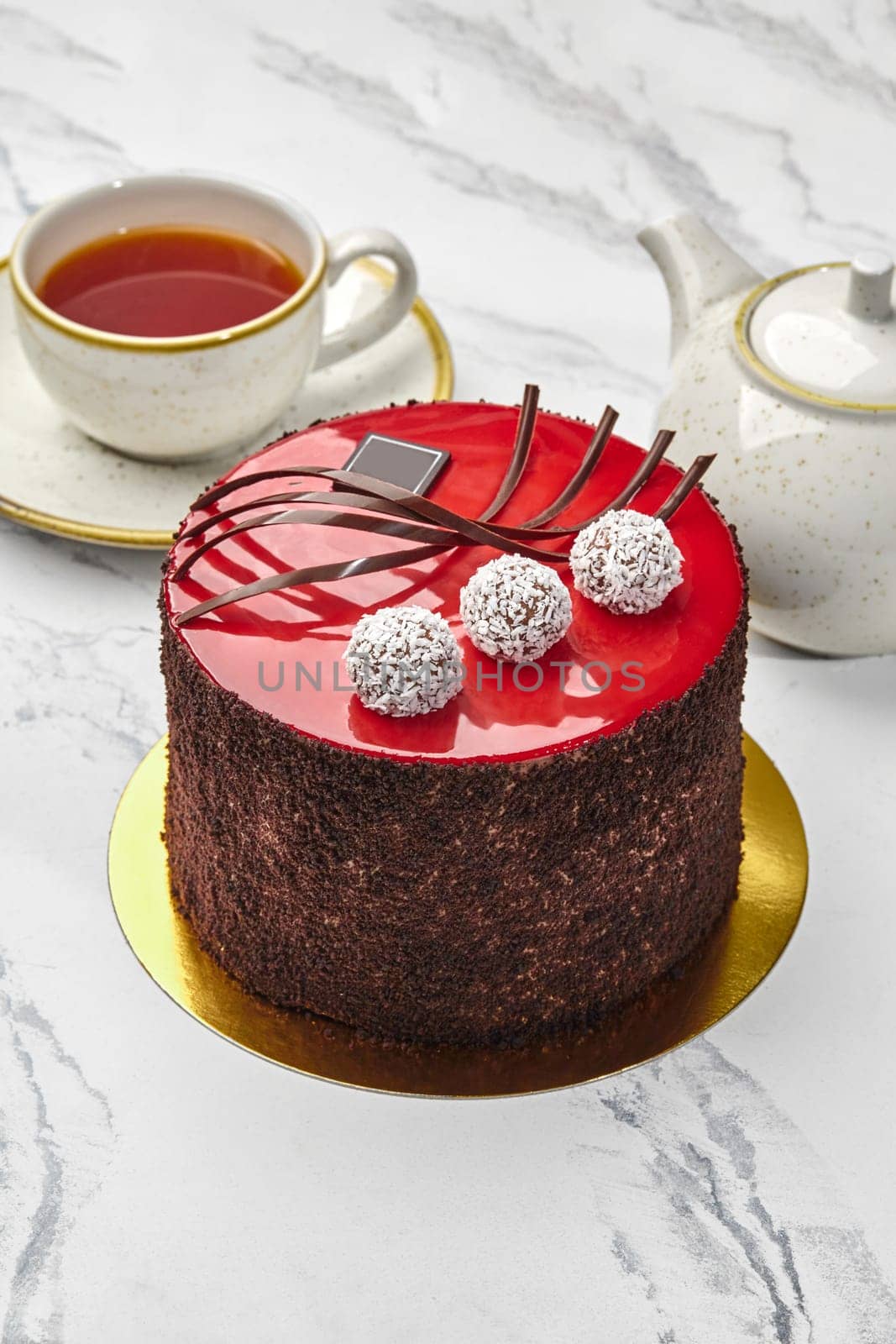 Inviting tea setting with striking red glazed chocolate cake decorated with coconut truffles, teacup, and teapot on marble background