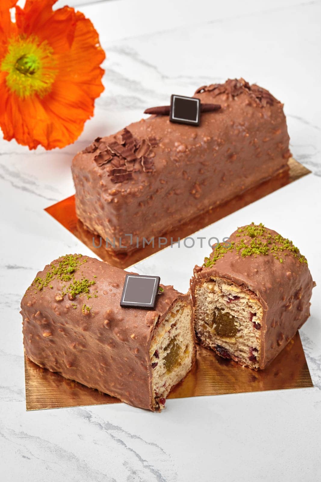 Handcrafted chocolate glazed loaf cake with fruit filling, berries and nuts, sprinkled with pistachio crumbs for delicious snacking, presented on marble surface with vibrant orange poppy flower