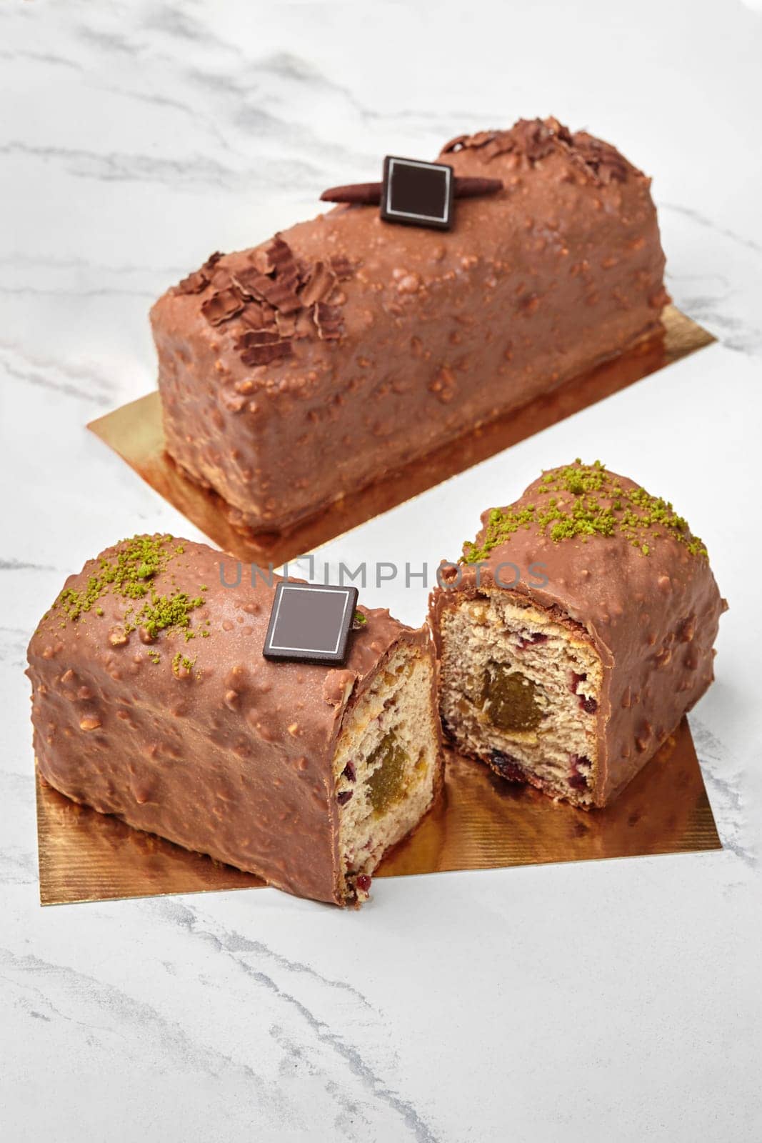 Sliced artisanal loaf cake with fruit jam filling and dried berries, coated with milk chocolate and nuts glaze, decorated with dark chocolate branding plaque and pistachio crumble on golden cardboard