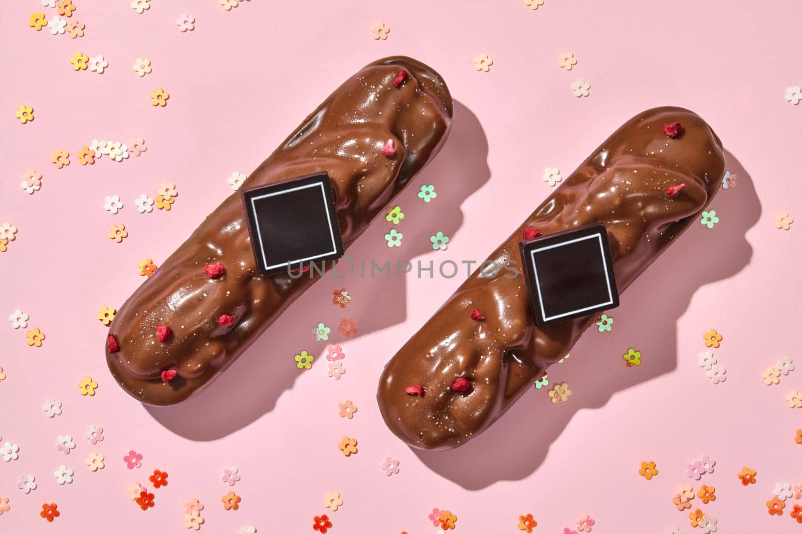 Top view of two milk chocolate-covered pecan bars, sprinkled with dried berry pieces, elegantly presented on pink background with scattered colorful flowers shaped confetti