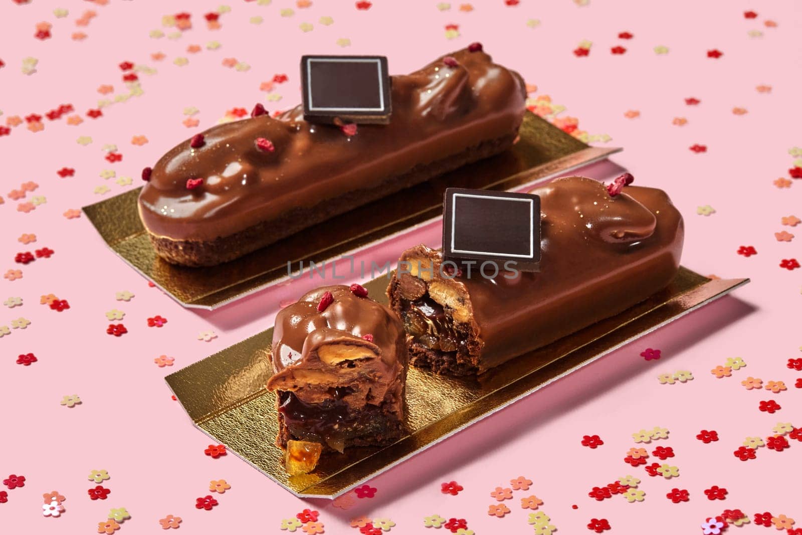 Delicious streusel-based bars with caramelized fruits and pecans, coated in milk chocolate, adorned with dried berries on pink background with scattered flower shaped confetti. Artisan handmade sweets