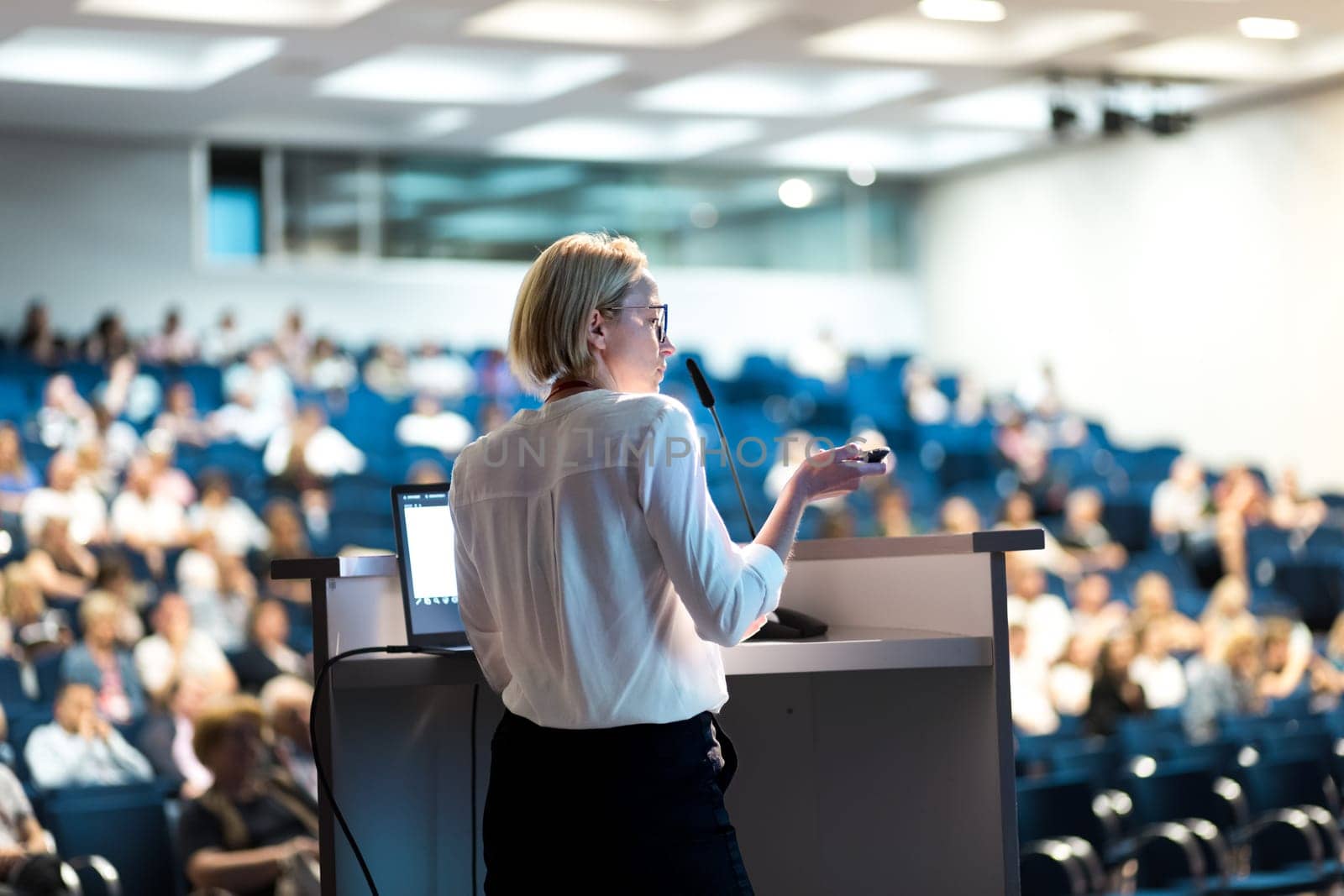 Female speaker giving a talk on corporate business conference. Unrecognizable people in audience at conference hall. Business and Entrepreneurship event