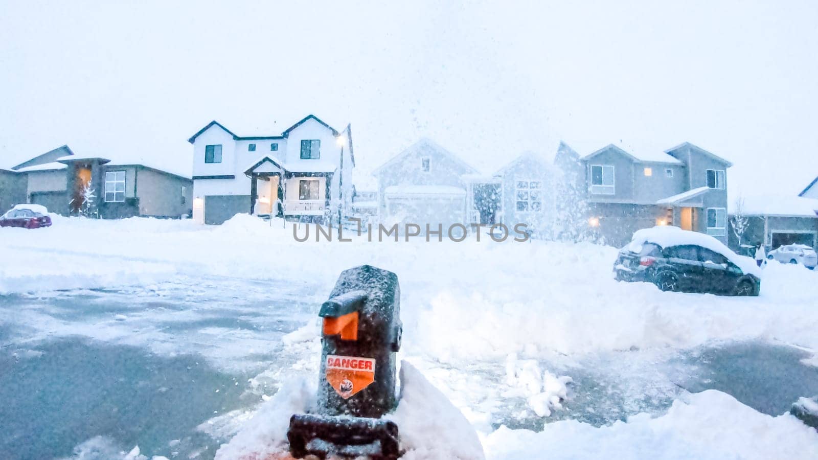 Denver, Colorado, USA-February 4, 2024-Snow blower is at work in the early evening light, clearing a path in a neighborhood where houses and a large snow pile are softly illuminated against the dusky sky.