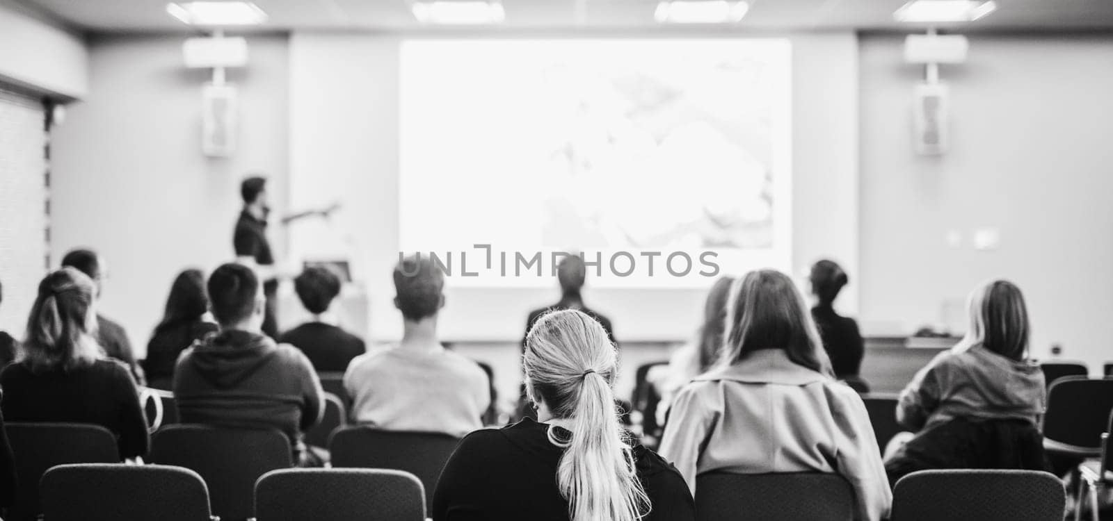 Speaker giving a talk in conference hall at business event. Rear view of unrecognizable people in audience at the conference hall. Business and entrepreneurship concept. Blue tones black and white image