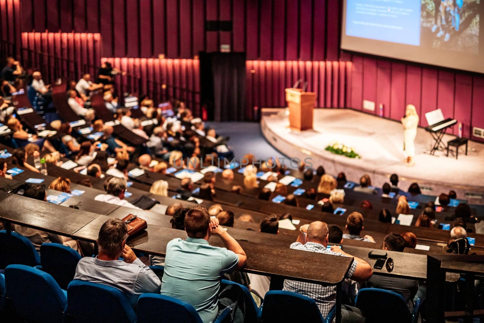 Business and entrepreneurship symposium. Female speaker giving a talk at business meeting. Audience in conference hall. Rear view of unrecognized participant in audience.