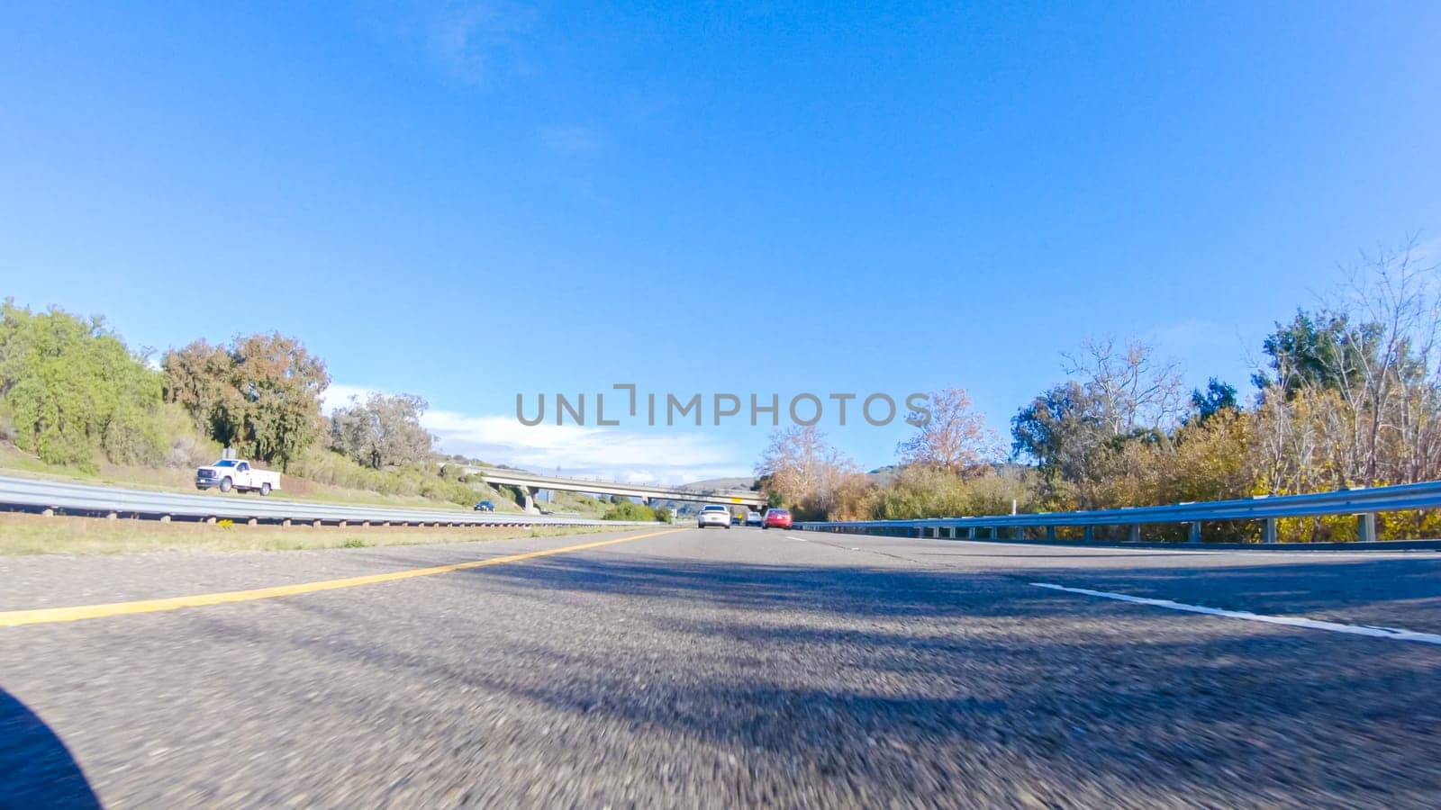 On a crisp winter day, a car cruises along the iconic Highway 1 near San Luis Obispo, California. The surrounding landscape is brownish and subdued, with rolling hills and patches of coastal vegetation flanking the winding road.