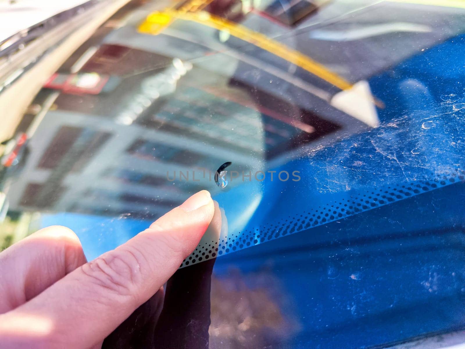 A hand points to a small chip on the driver-side window of a parked car, highlighted in bright daylight