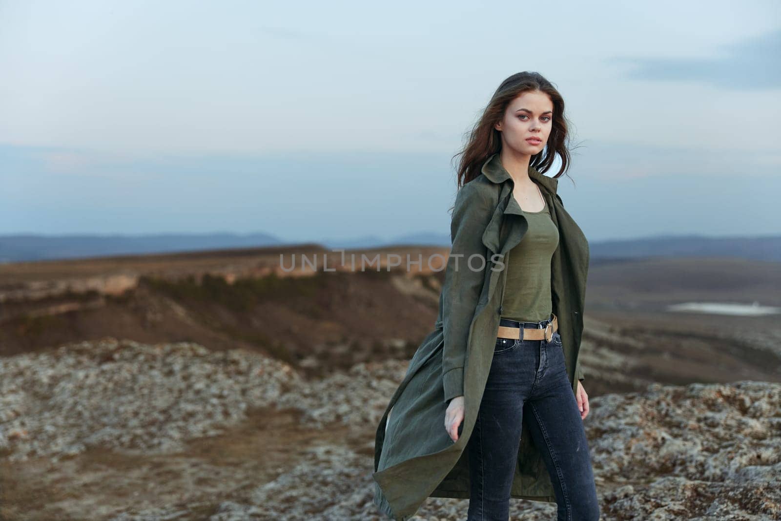 Mysterious woman in trench coat gazes at majestic mountains from hilltop perch