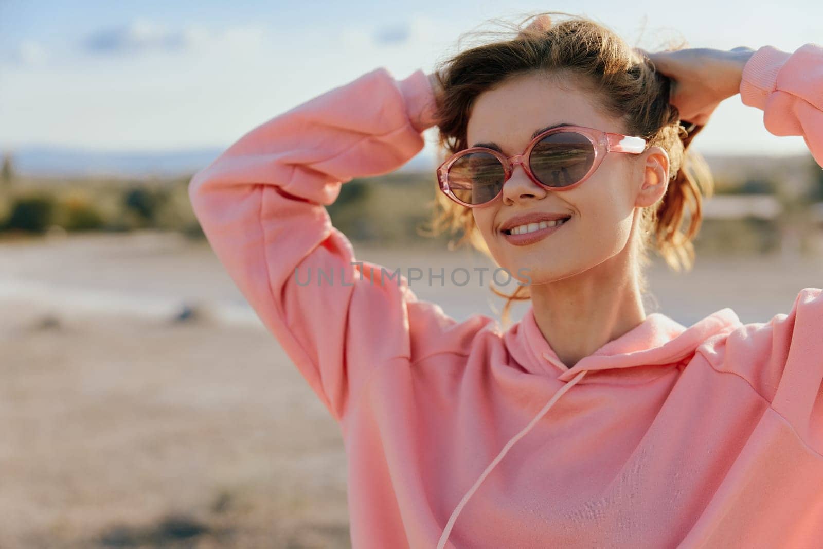 Fashionable woman in sunglasses and pink sweatshirt posing with hands on head in desert landscape by Vichizh