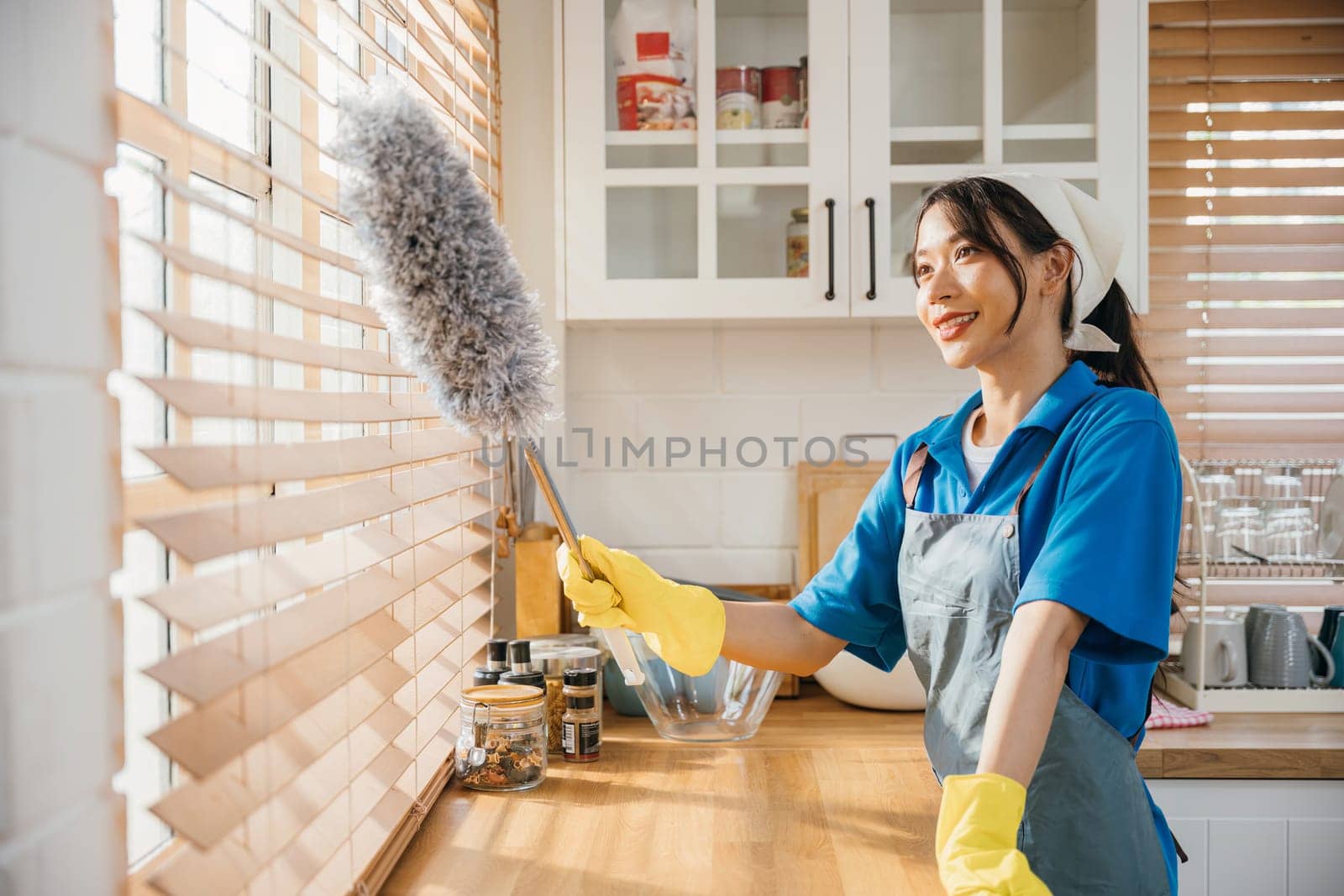 Portrait of a smiling Asian woman cleaning window blinds. She stands duster in hand enjoying her routine housework. Modern cleaning occupation ensuring hygiene and cleanliness. whisk