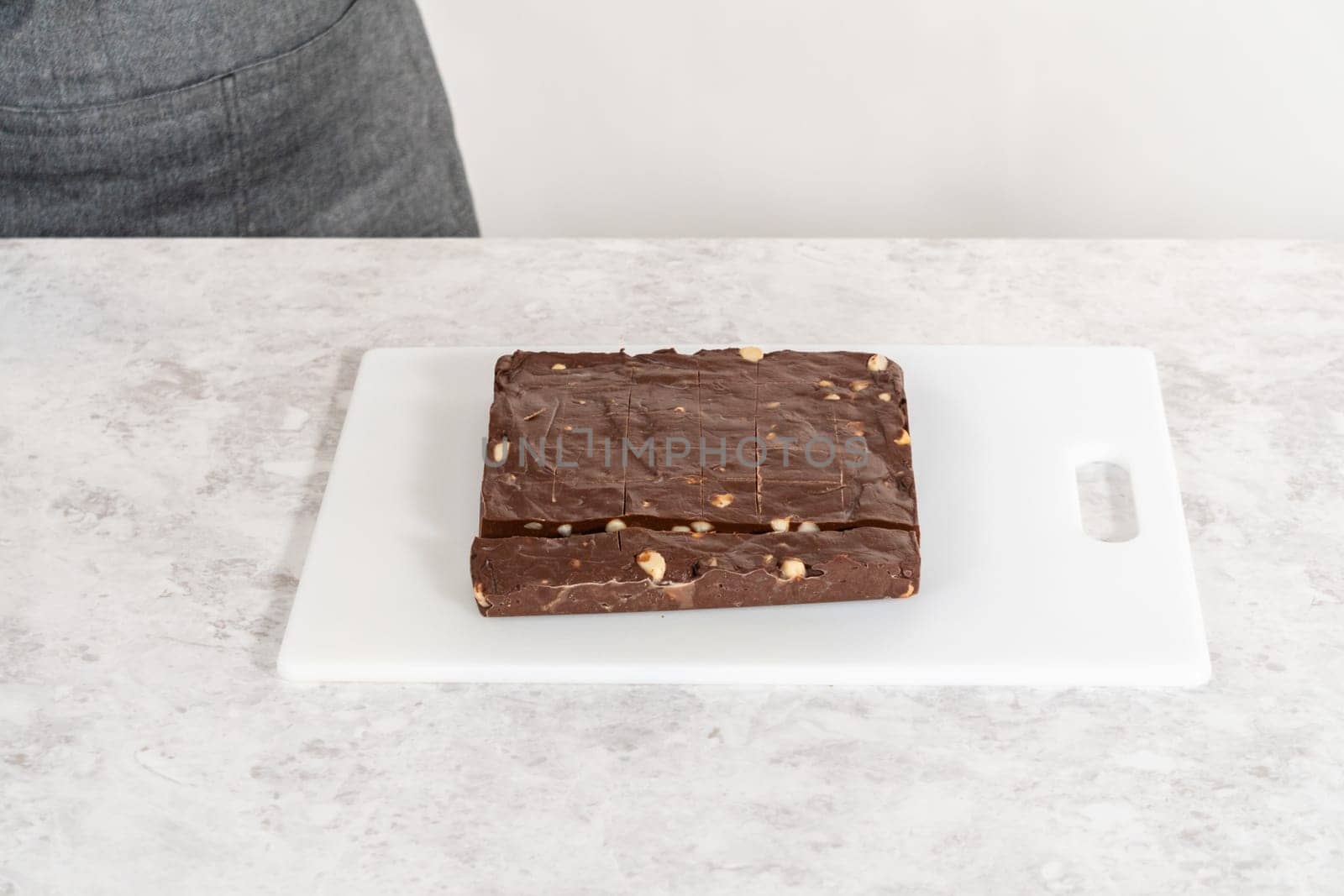 Cutting chocolate macadamia fudge with a large kitchen knife into square pieces on a white cutting board.