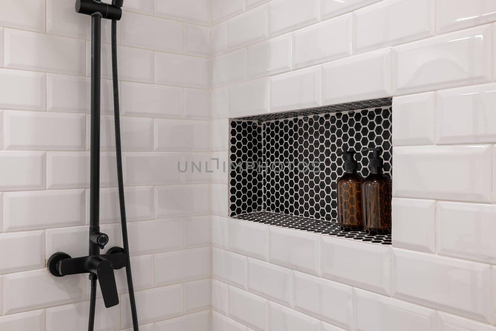 Elegant white tiled shower with black fixtures, featuring a built-in shelf with stylish black tiles and brown soap bottles. Perfect for contemporary bathroom designs.