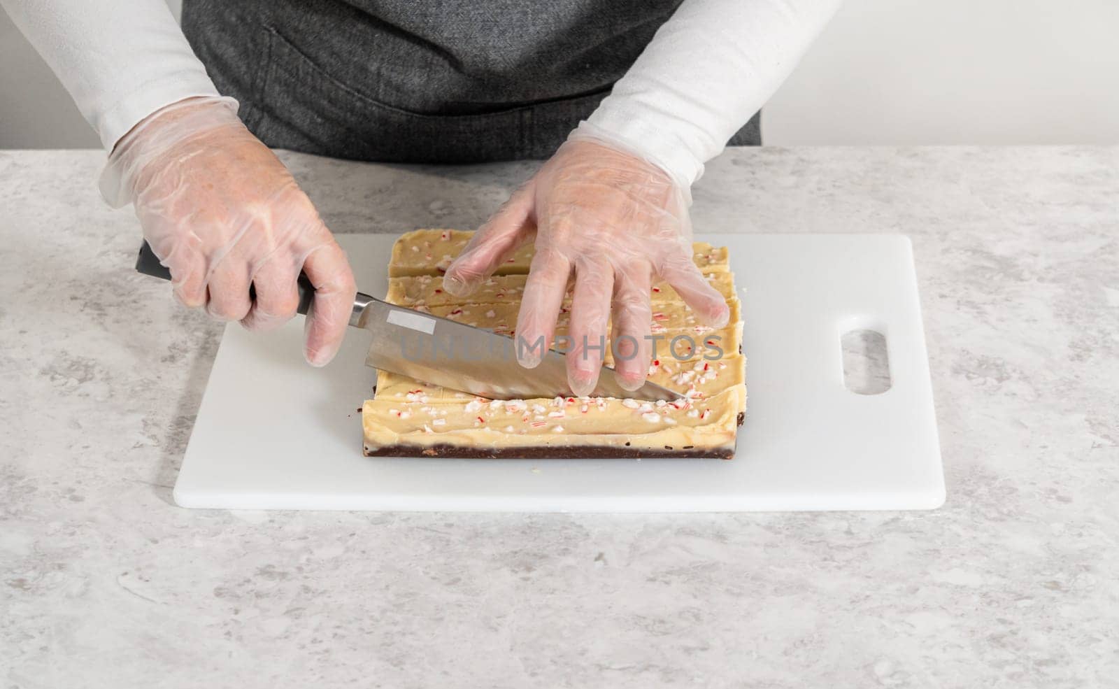 Cutting candy cane fudge with a large kitchen knife into square pieces on a white cutting board.