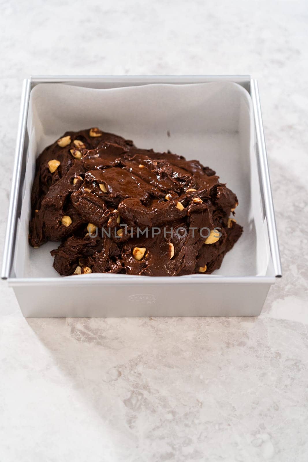 Filling square cheesecake pan lined with parchment paper with fudge mixture to prepare chocolate macadamia fudge.