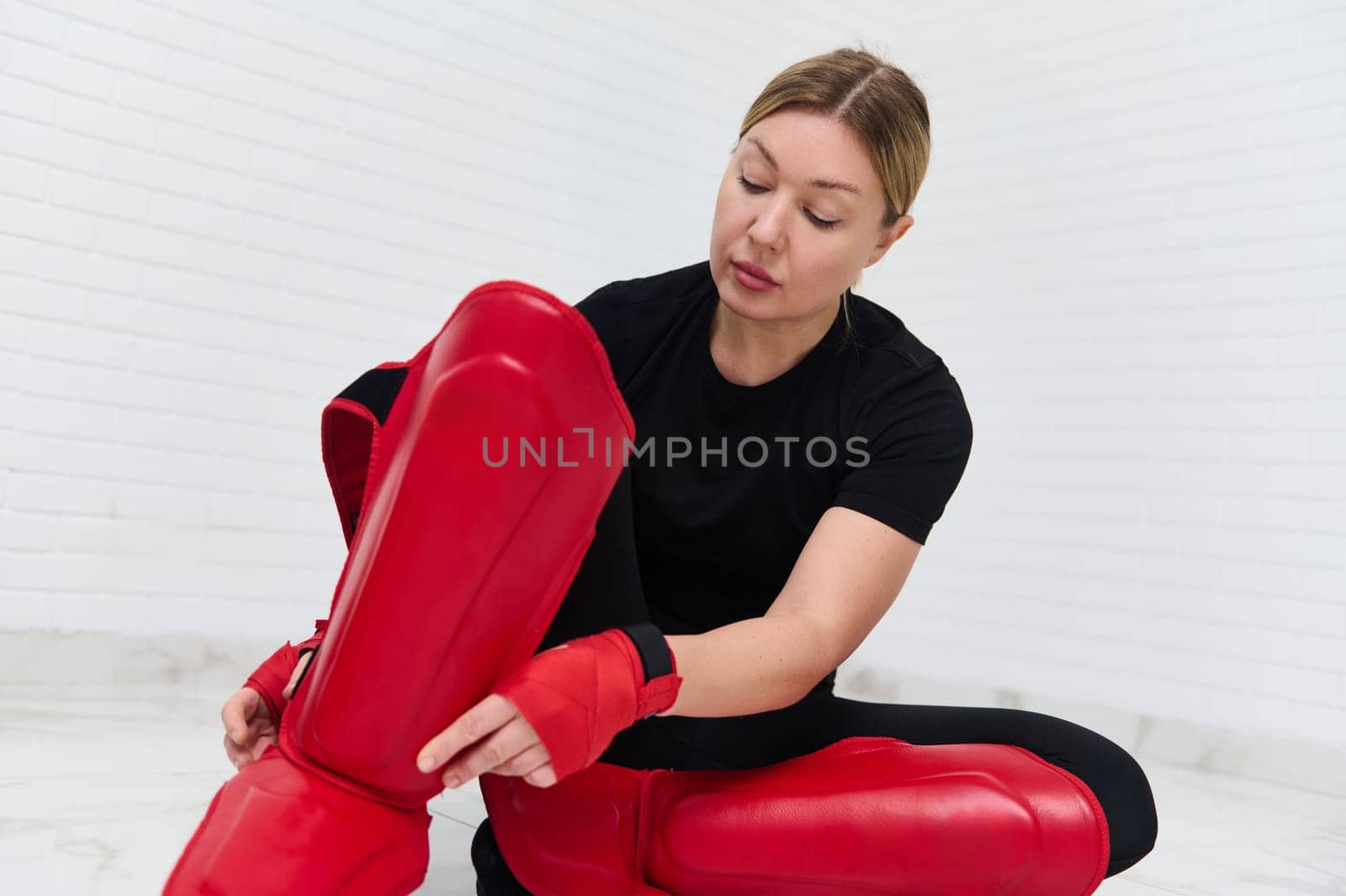 Young European woman fighter, kickboxer putting on red protective kickboxing equipment on legs, ready for training, isolated over white background. People. Kickboxing workout. Martial art. Challenge