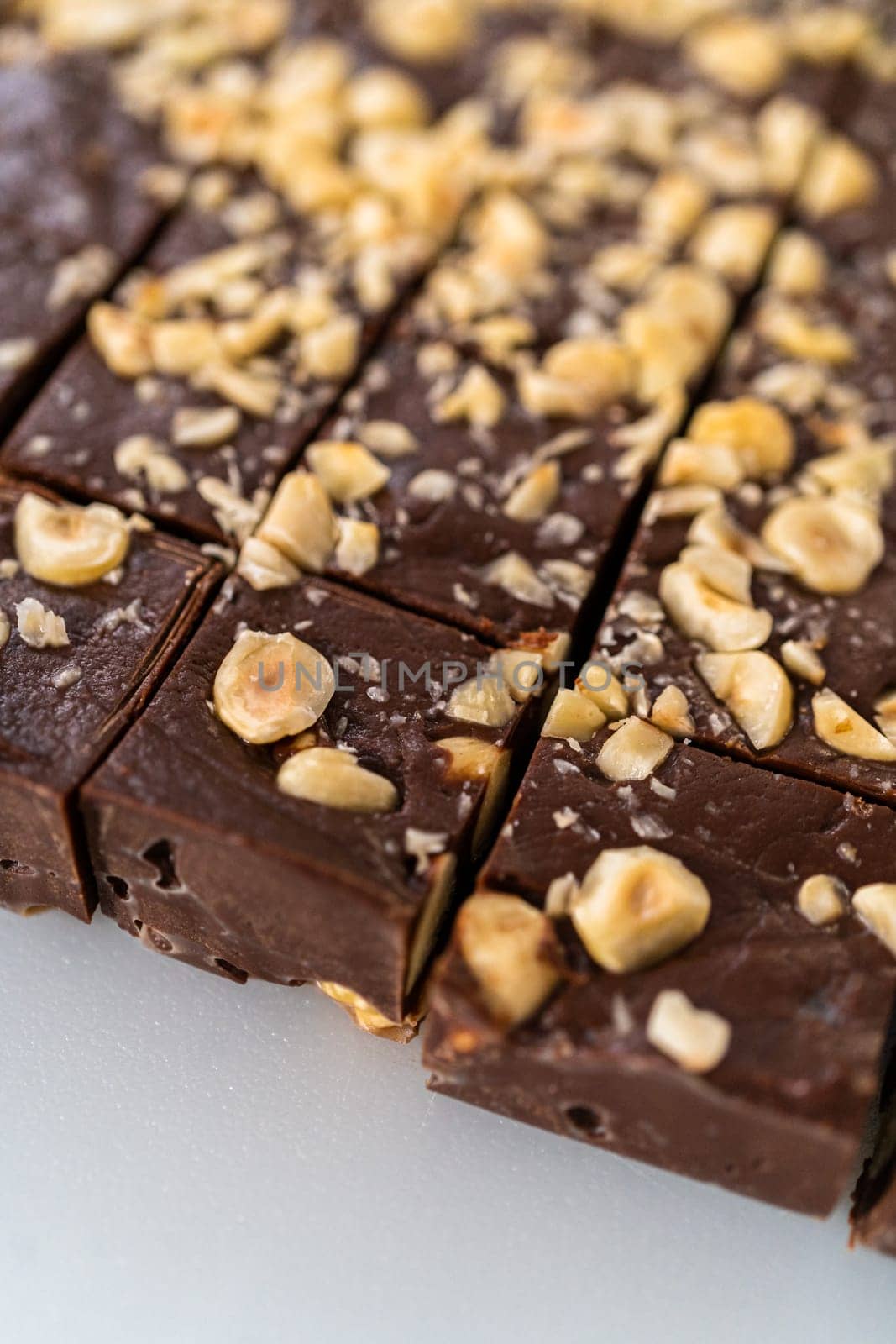 Cutting chocolate hazelnut fudge with a large kitchen knife into square pieces on a white cutting board.