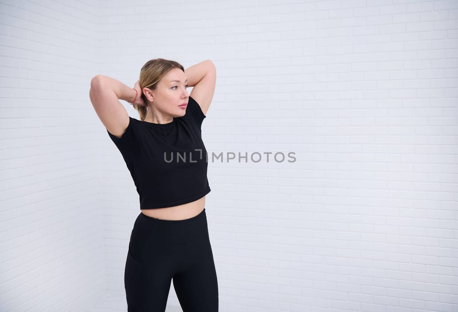 Beautiful Caucasian European athletic woman 30s in black sportswear, tying a ponytail, isolated over white wall background